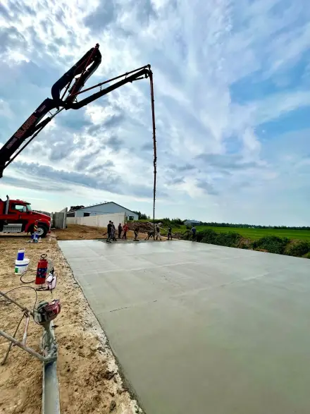 Grande dalle de béton fraîchement coulée, une équipe à la truelle au fond et une pompe à béton déployée.