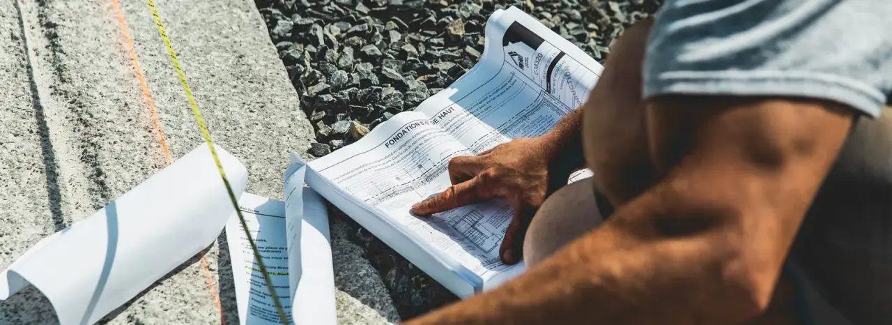 Deux mains posées sur un plan intitulé « Fondation vue de haut », déroulé sur une dalle de béton à côté d’un ruban à mesurer et d’une lettre à en-tête Bâti-Forme.