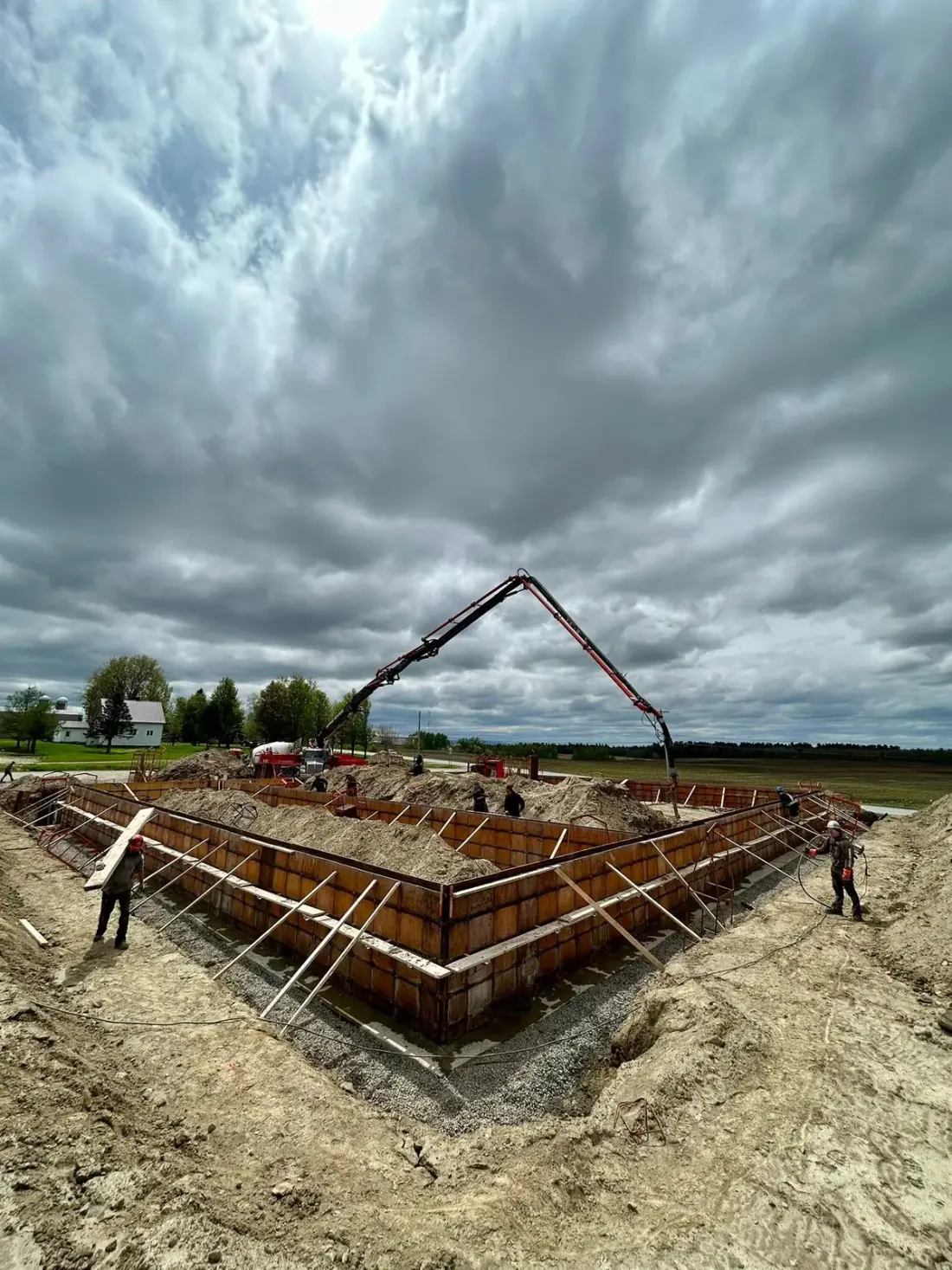 Périmètre de fondation entièrement coffré au fond d’une excavation, la flèche d’une pompe à béton déployée au-dessus, des ouvriers au travail.