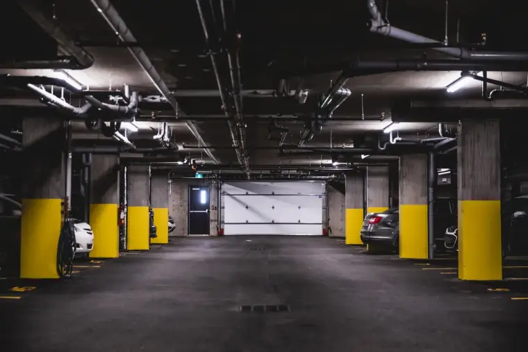 Intérieur d’un stationnement souterrain : colonnes et dalles de béton, tuyauterie apparente au plafond.