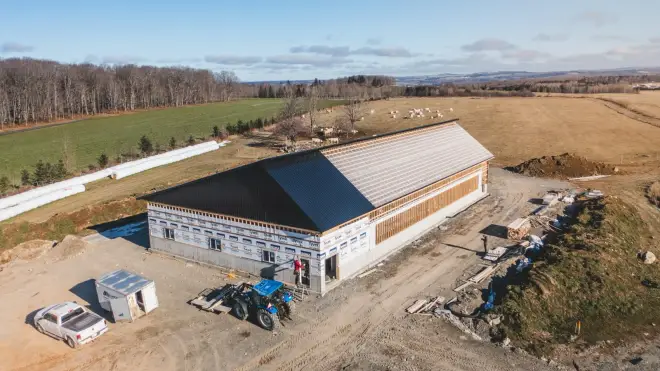Vue aérienne d’un long bâtiment de ferme en construction, assis sur un mur de fondation de béton, champs et troupeau autour.